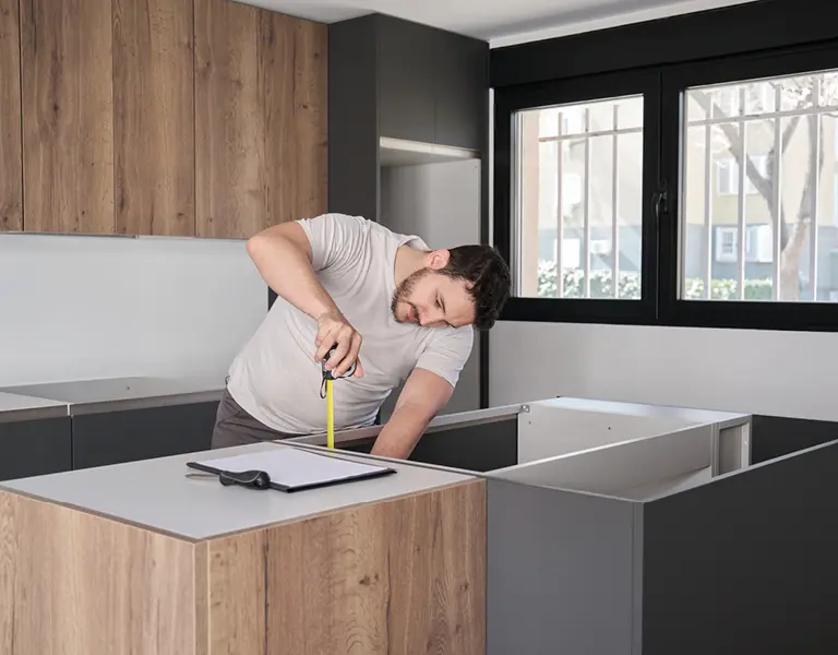 Handwerker misst eine Arbeitsplatte in einer modernen Küche mit Holzfronten und grauen Schränken, im Hintergrund große Fenster mit schwarzem Rahmen.